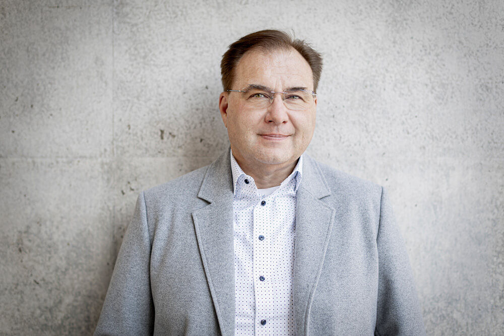 Mann in grauem Sakko steht vor Betonwand und lächelt, Businessfoto von Iris Schmitz